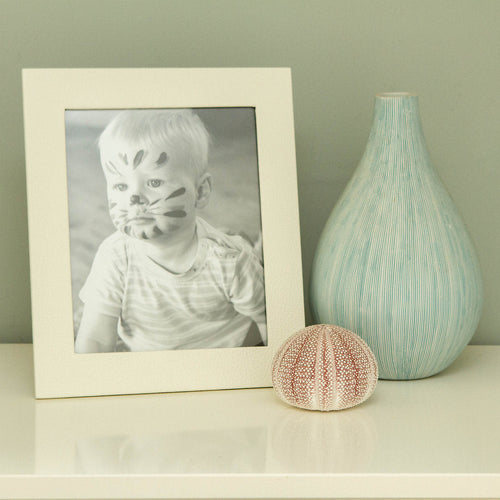 Ivory Landscape Leather Frame 7x5 displaying a black and white photo of a child with face paint next to a decorative vase and shell.